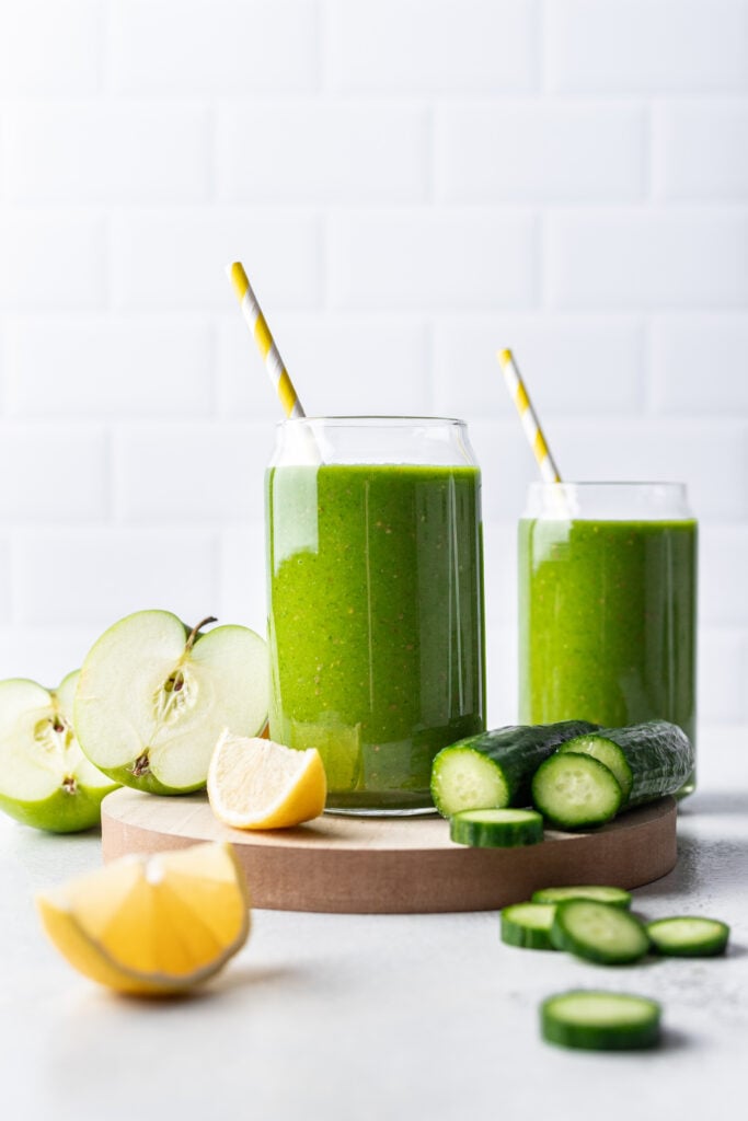 green smoothie in 2 glasses with apple, lemon and cucumber around it