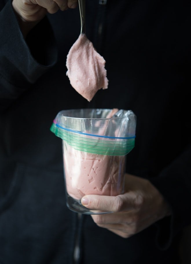 Hand holding a glass with a plastic bag full of pink frosting inserted and folded over the edges with a spoon dropping more frosting inside the bag