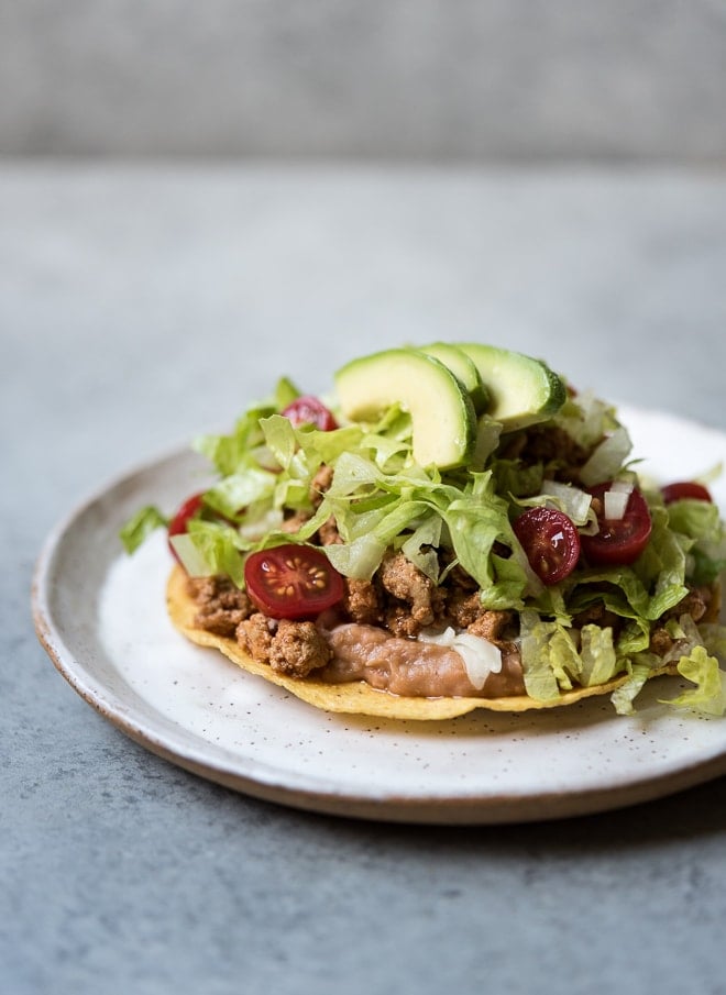 refried beans, ground turkey, lettuce, tomato and avocado on a crispy tostada on a white plate