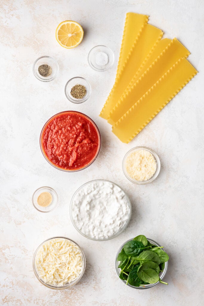 ingredients in a bowls like tomato sauce, cottage cheese, lasagna noodles, spices, spinach