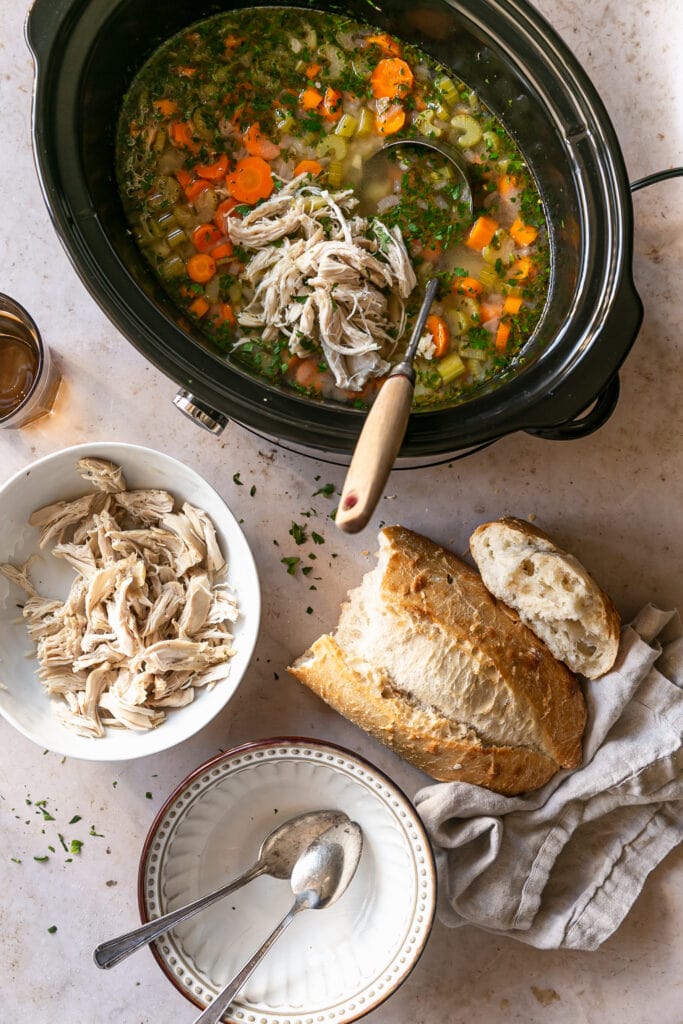 Slow cooker on countertop with shredded chicken, crusty bread, bowls and spoons