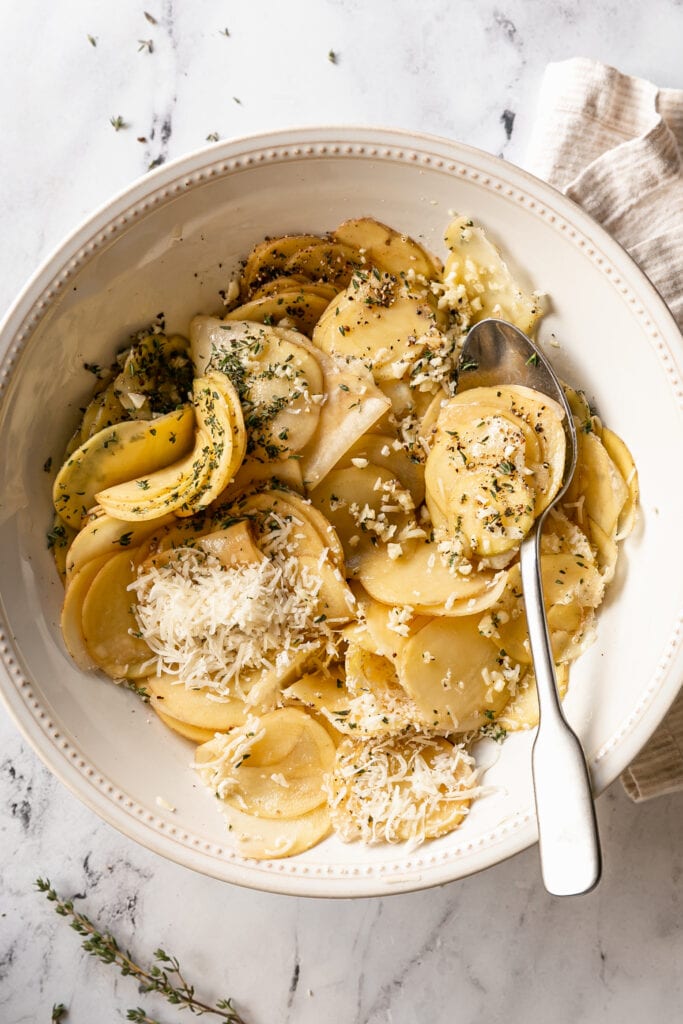 sliced potatoes in a bowl with garlic, thyme, and parmesan cheese