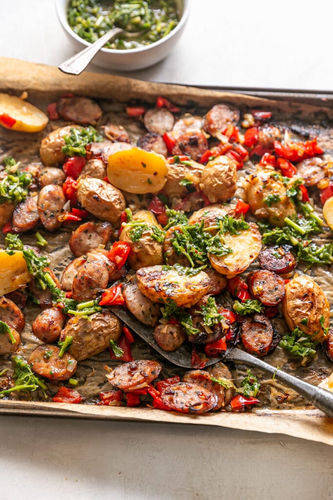 LIBAD Sheet Pan Sausage and Potatoes - cooked sheet pan with spatula, sausage, potatoes and peppers with chimichurri
