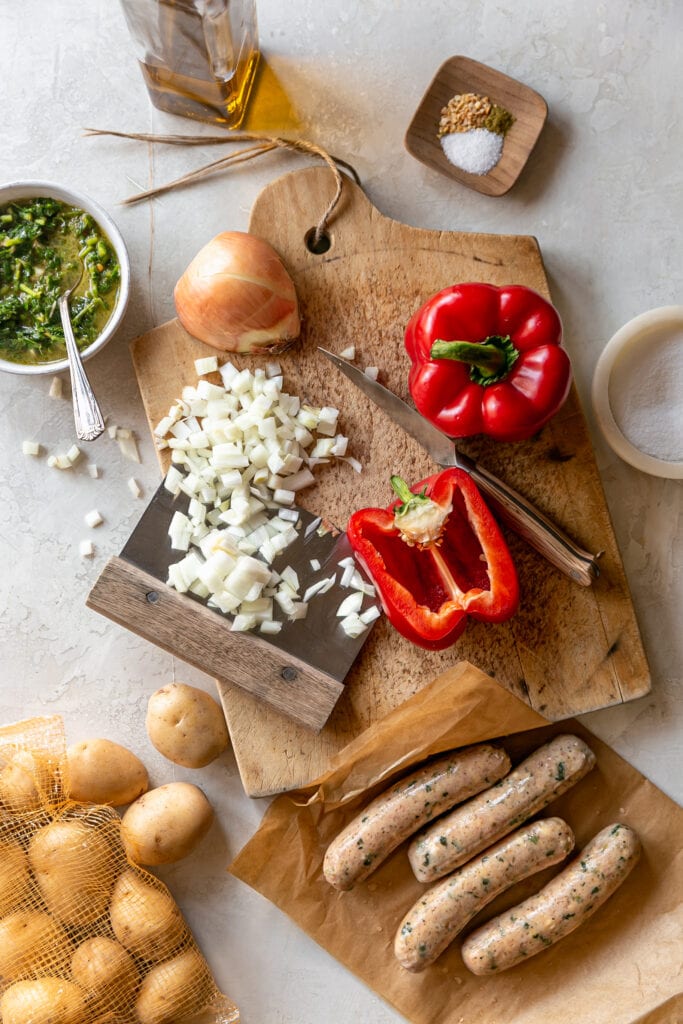 Sheet Pan Sausage and Potatoes with onion and red bell pepper on a cutting board with knife, baby potatoes, and chimichurri