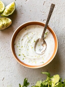 Easy 4-Ingredient Lime Crema - finished lime crema in orange bowl with spoon, limes and cilantro