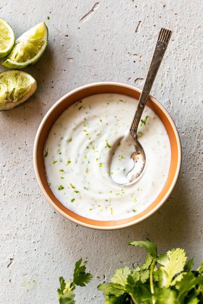Easy 4-Ingredient Lime Crema - finished lime crema in orange bowl with spoon, limes and cilantro
