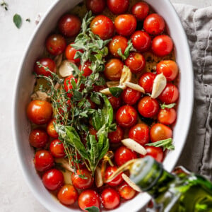 Quick Tomato Garlic Confit - cherry tomatoes, herbs and garlic in baking dish with olive oil pouring in