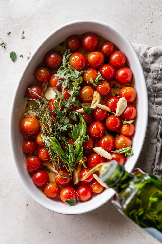 Quick Tomato Garlic Confit - cherry tomatoes, herbs and garlic in baking dish with olive oil pouring in