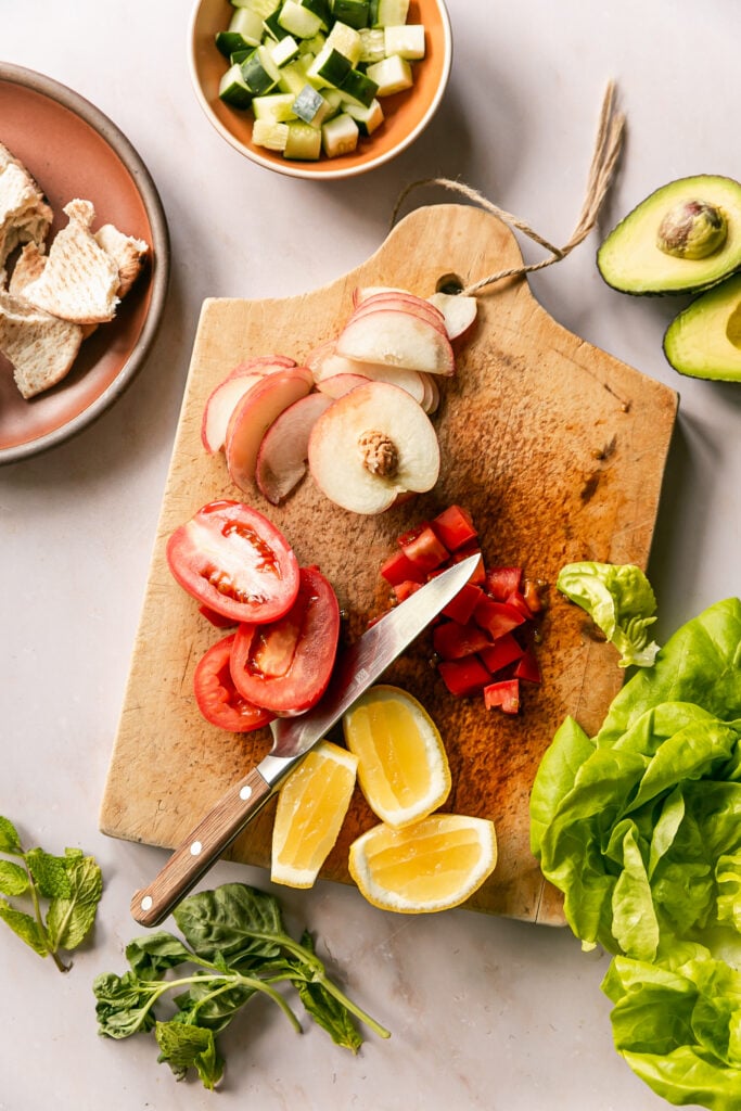 Summer Fattoush Salad - slicing lemon, tomato, peach on cutting board with lettuce, tomato, cucumber and pita