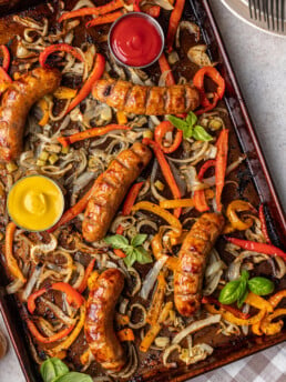 peppers, onions and sausage on a sheet pan with mustard and ketchup in small bowls