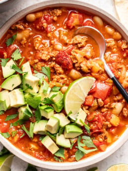 bowl of chicken chili with a spoon and topped with avocado and a lime