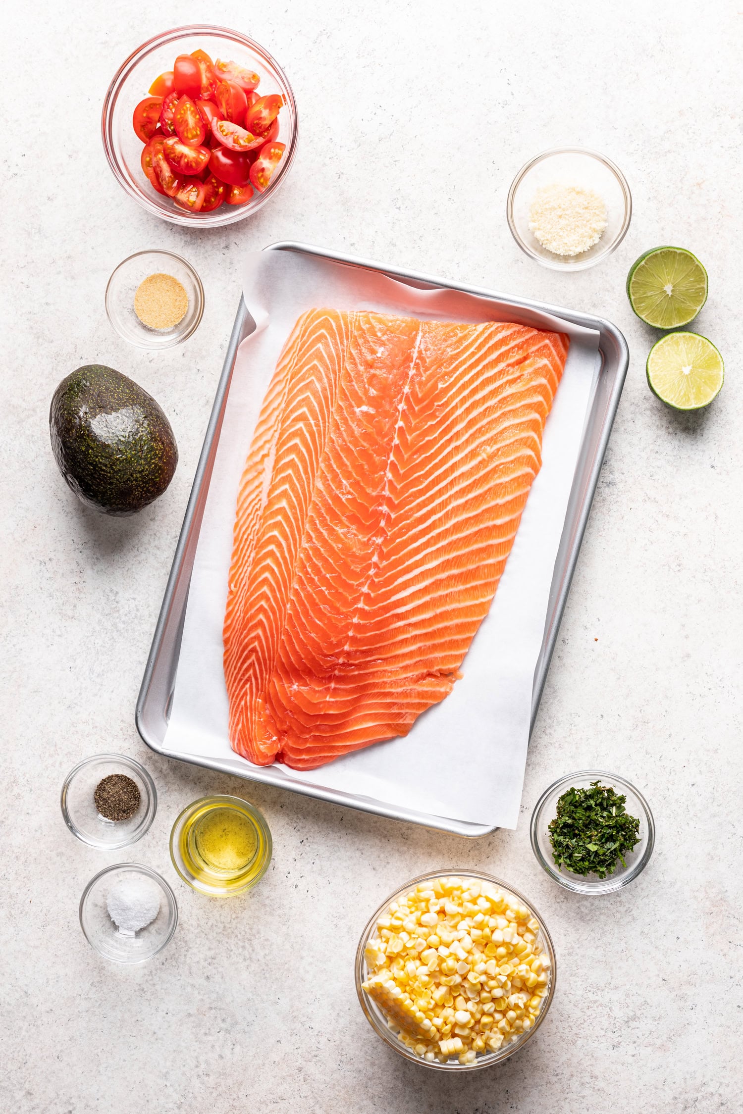 large piece of salmon on a baking sheet with bowls of ingredients surrounding