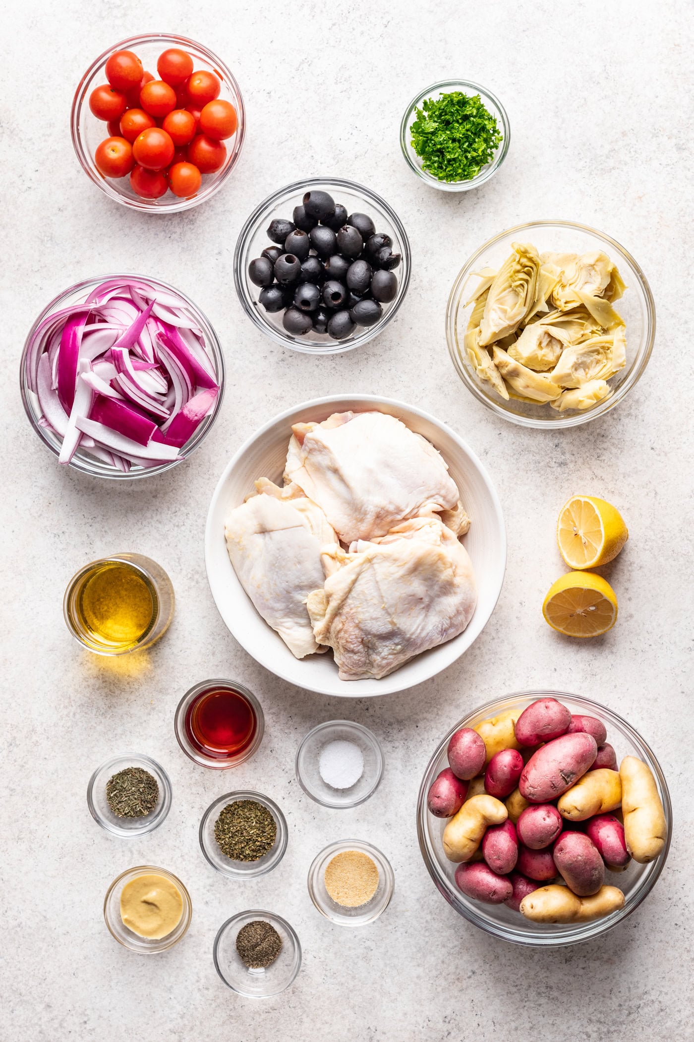 bowls of ingredients on a grey background such as raw chicken thighs, olives, onion and potatoes