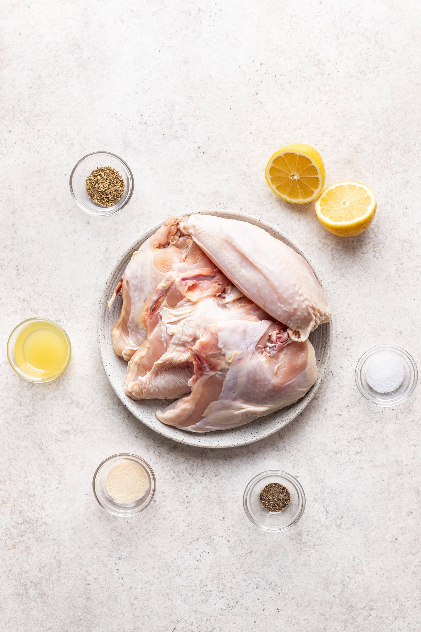 A plate with raw chicken pieces, ready to become lemon roasted chicken, is surrounded by bowls of black pepper, salt, garlic powder, lemon juice, olive oil, and two lemon halves on a light-colored surface.