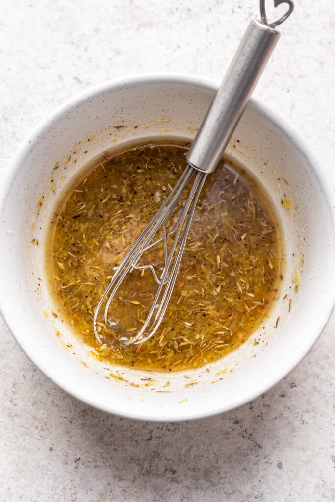 A metal whisk rests in a white bowl filled with a yellowish herb-infused vinaigrette, perfect for drizzling over lemon roasted chicken, on a light stone surface.