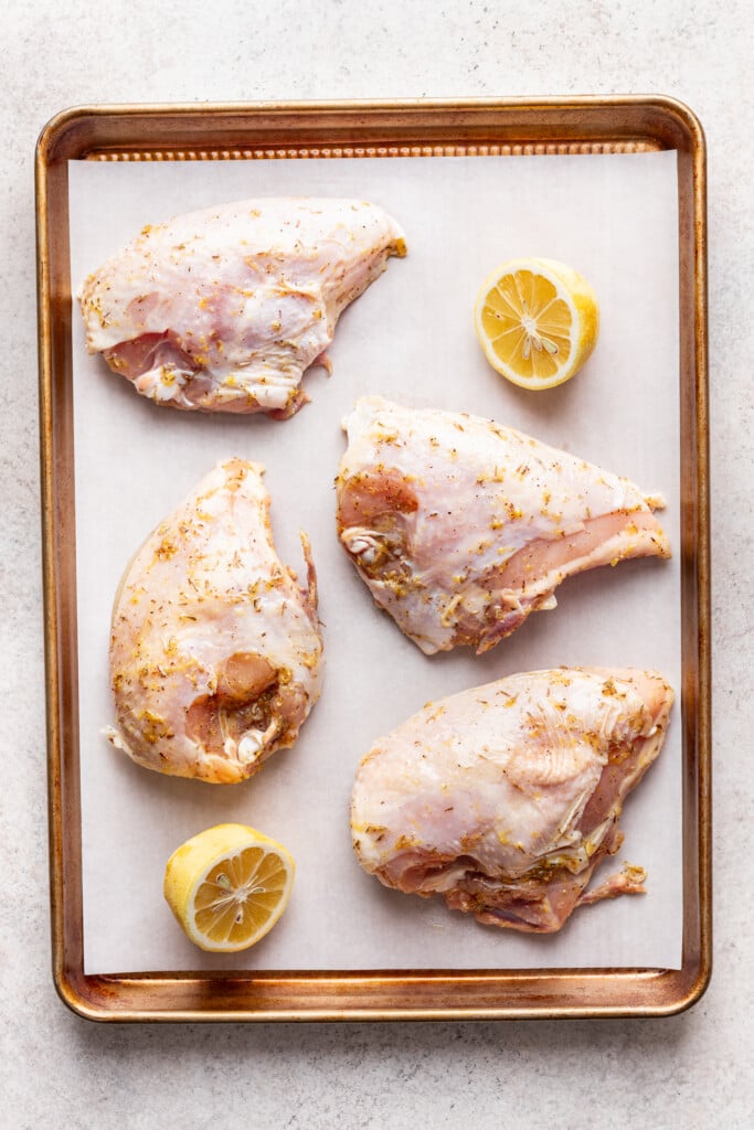Four raw, seasoned chicken breasts are arranged on a parchment-lined baking sheet, with two lemon halves nestled among them for a classic lemon roasted chicken preparation.