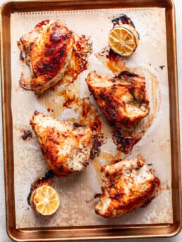 Four roasted bone-in chicken breasts with golden, crispy skin sit on a parchment-lined baking sheet. This lemon roasted chicken is paired with halved, roasted lemons, their juices and browned bits scattered around for extra flavor.
