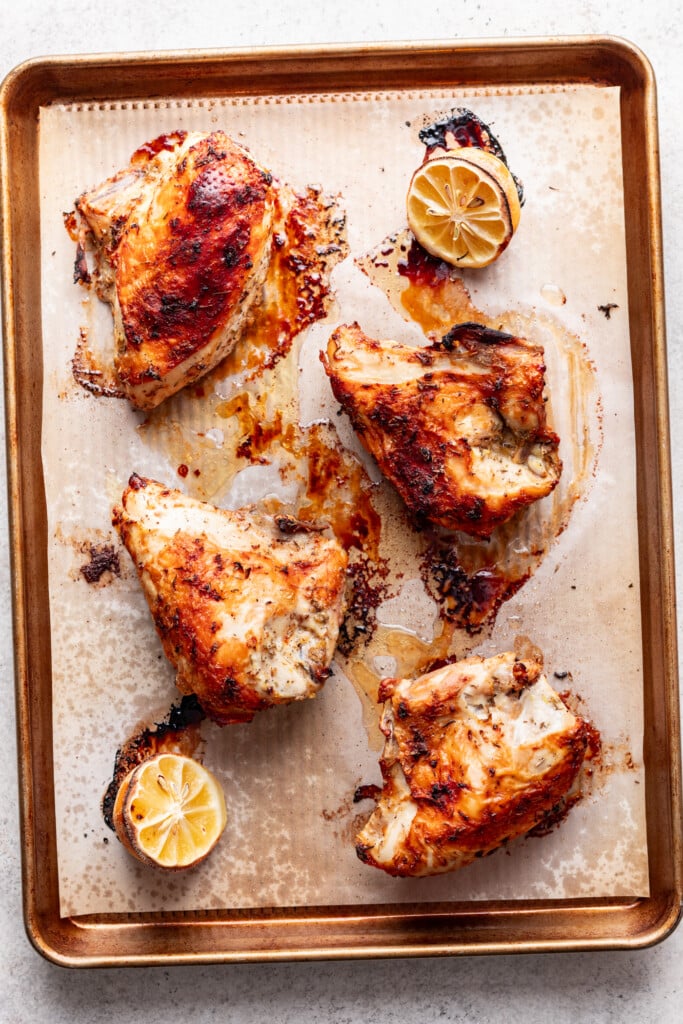 Four roasted bone-in chicken breasts with golden, crispy skin sit on a parchment-lined baking sheet. This lemon roasted chicken is paired with halved, roasted lemons, their juices and browned bits scattered around for extra flavor.