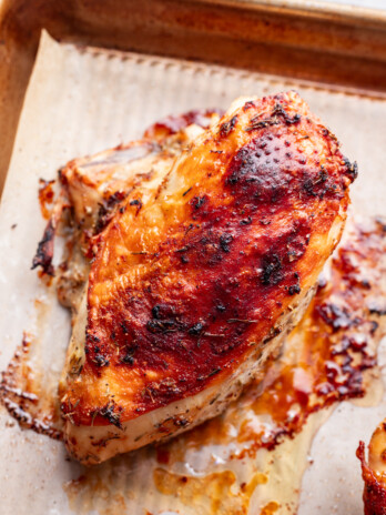 A piece of lemon roasted chicken breast with a browned, seasoned crust sits on parchment paper on a baking sheet, with juices and char marks visible.