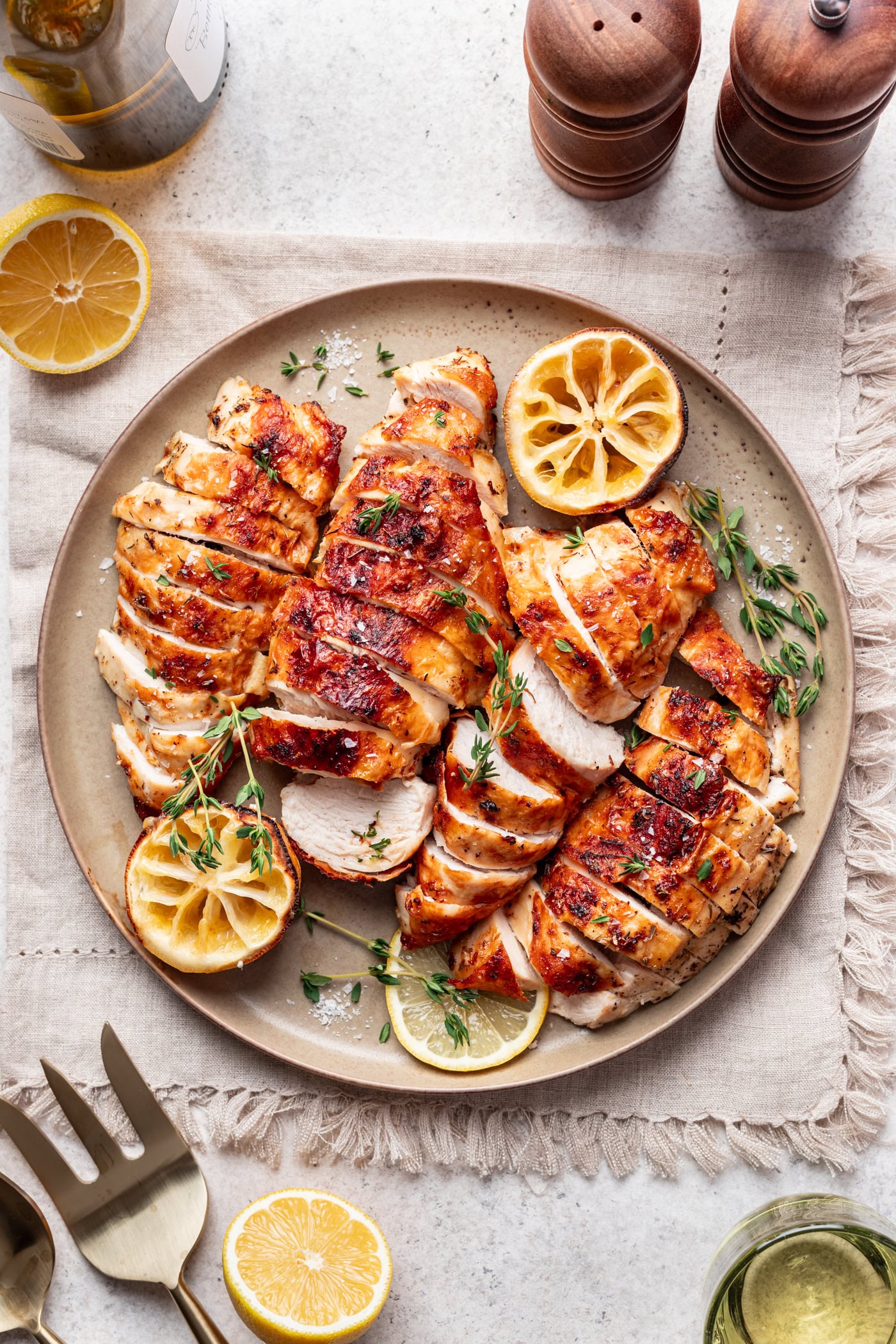 Sliced, lemon roasted chicken breast garnished with fresh thyme and lemon slices on a beige plate, surrounded by cut lemons, a metal serving fork, salt and pepper shakers, and a glass of white wine on a light table.