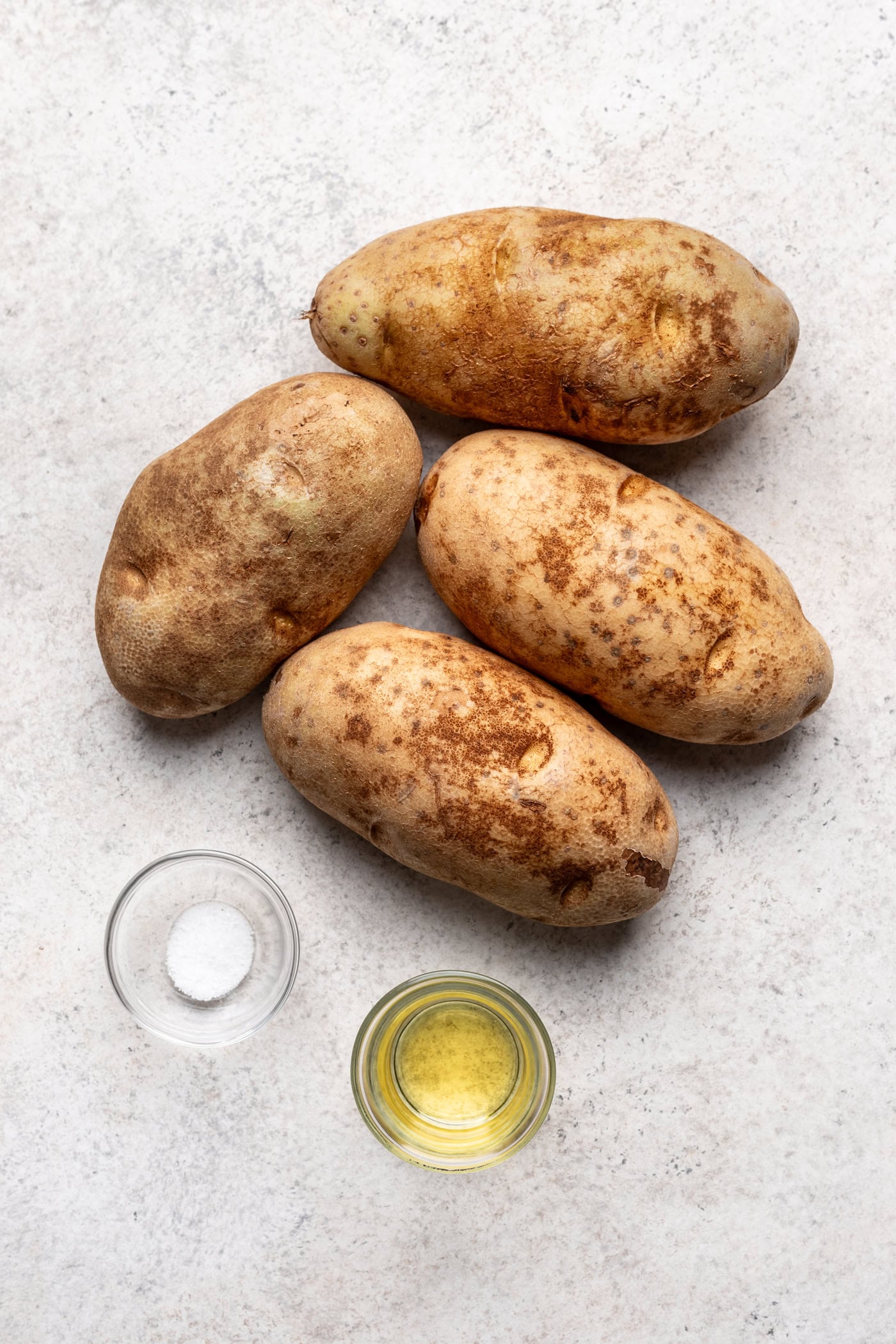 4 russet potatoes on a sheet pan with a small bowl of oil and salt