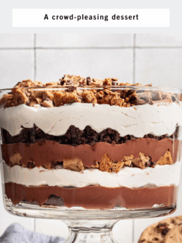 A layered trifle dessert with chocolate chip cookies, brownies, chocolate pudding, and whipped cream in a glass dish on a kitchen counter, with a plate of cookies and a napkin nearby. Text reads: Chocolate Chip Cookie Brownie Trifle.