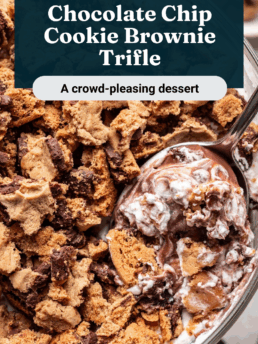 A glass bowl filled with chocolate chip cookie brownie trifle, topped with cookie pieces, whipped cream, and chocolate, with a spoon scooping some out. Text overlay says “Chocolate Chip Cookie Brownie Trifle.”.