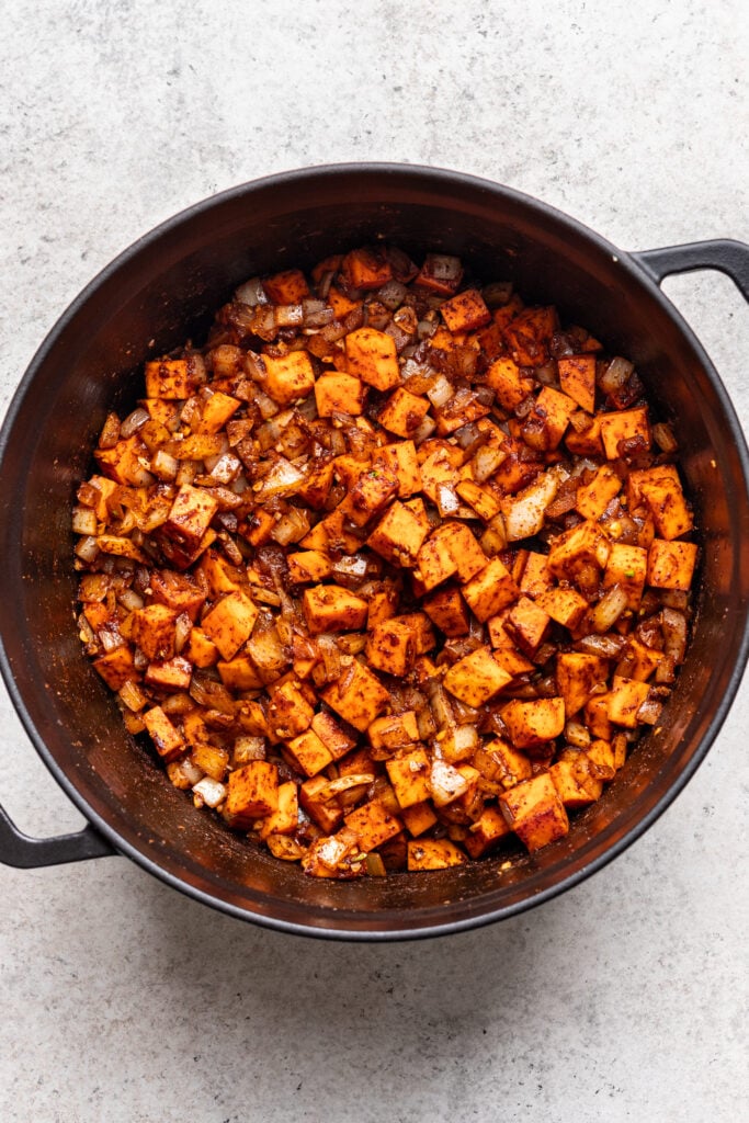 A black pot filled with sautéed diced sweet potatoes and chopped onions, seasoned with spices, sits on a light-colored countertop.