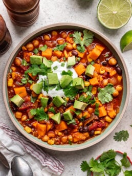 A bowl of vegetable chili topped with diced avocado, cilantro, and a dollop of sour cream. Ingredients like chickpeas, beans, corn, and tomato are visible. Lime halves, fresh cilantro, and pepper grinders are nearby.