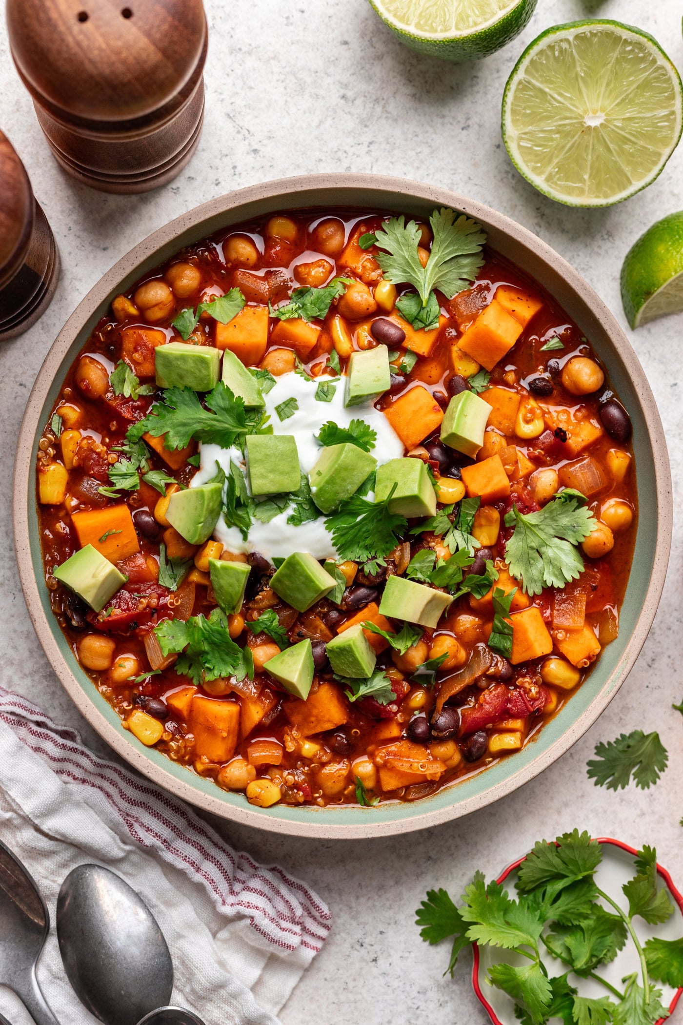 A bowl of vegetable chili topped with diced avocado, cilantro, and a dollop of sour cream. Ingredients like chickpeas, beans, corn, and tomato are visible. Lime halves, fresh cilantro, and pepper grinders are nearby.