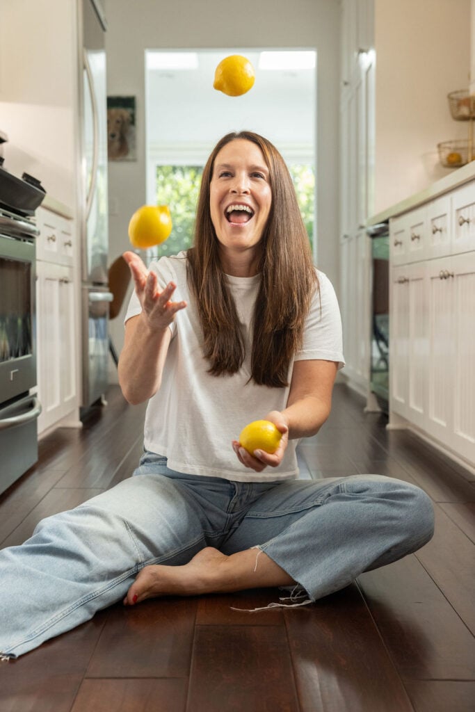 A woman sits cross-legged on a kitchen floor, smiling and juggling three lemons. She wears a white t-shirt and jeans, with bright light coming from a window behind her.