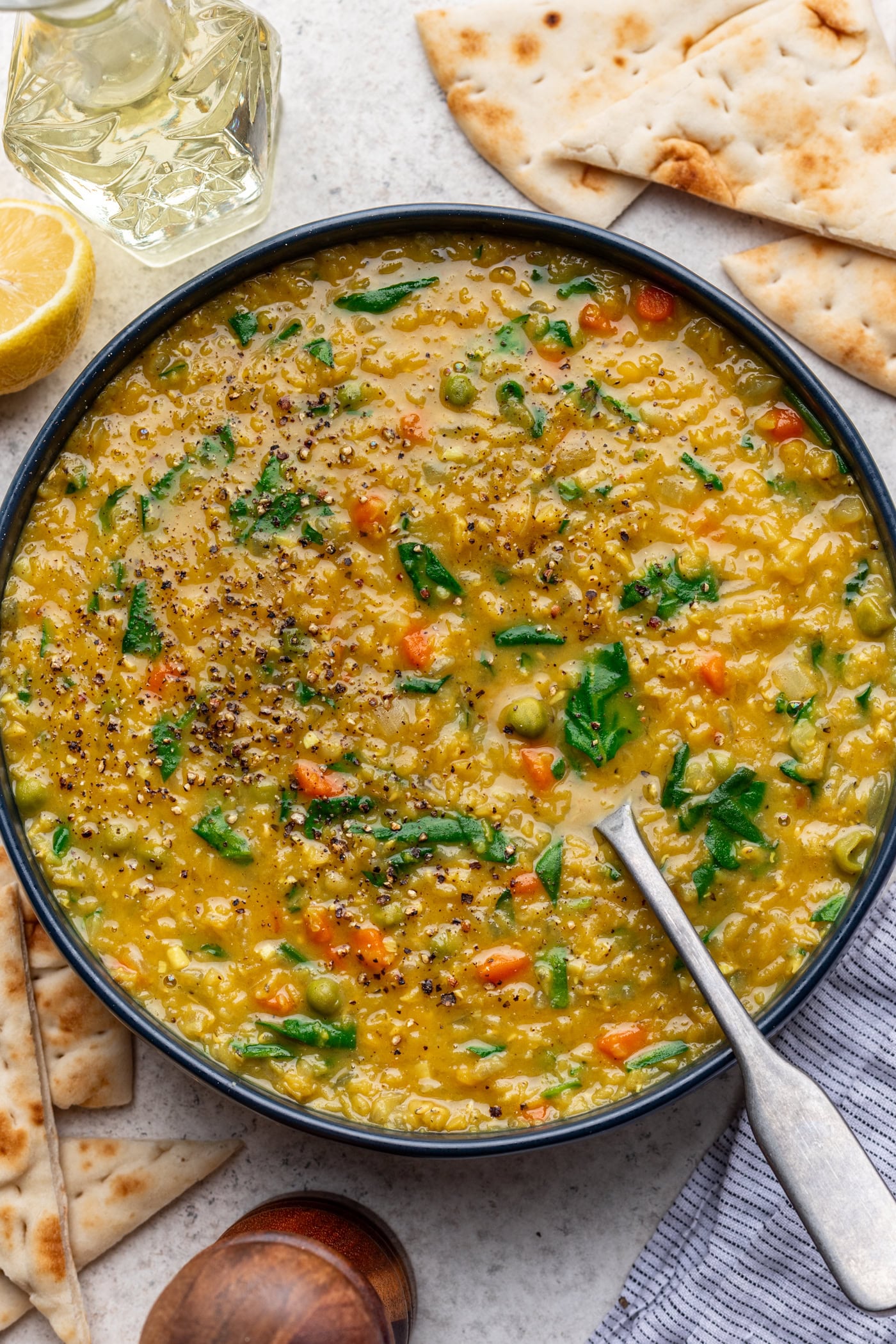 A bowl of yellow lentil soup with carrots, peas, and spinach, topped with cracked black pepper. Slices of flatbread, a lemon wedge, and a glass of water are nearby on a light surface. A spoon rests in the bowl.