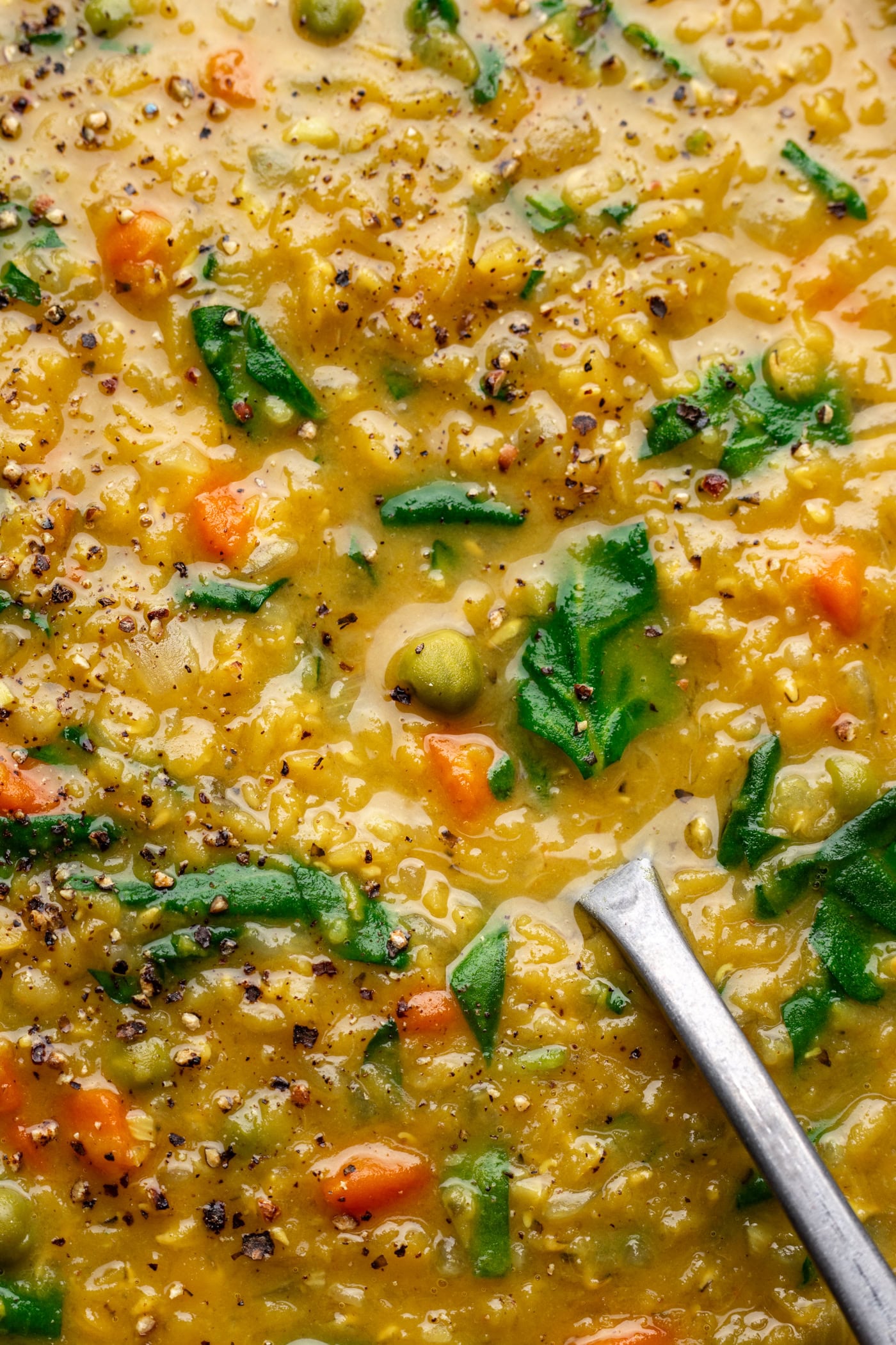 Close-up of a creamy lentil soup with visible pieces of carrot, spinach, green peas, and cracked black pepper, with a spoon partially submerged in the soup.