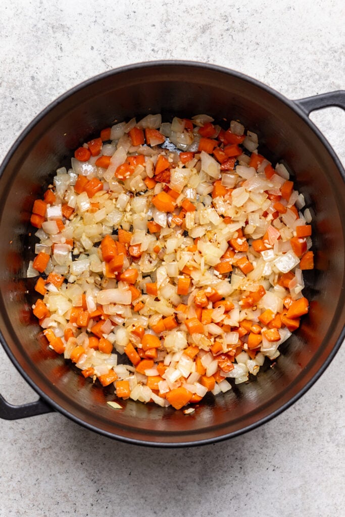 Chopped onions and carrots are sautéing in a large, black pot, with both vegetables softened and slightly browned. The pot sits on a light-colored surface.