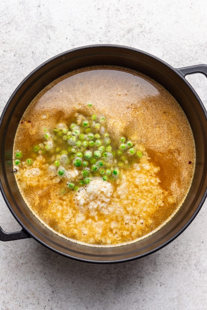 A black pot filled with broth, rice, green peas, and a dollop of cream or cheese sits on a light-colored surface, ingredients simmering together.