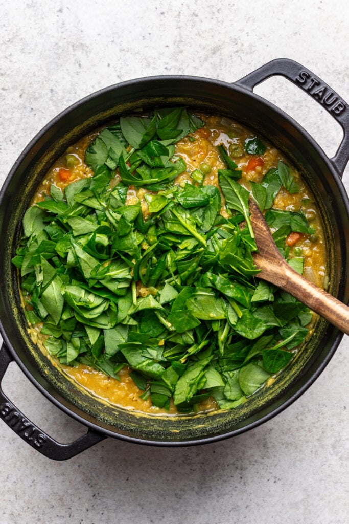 A black pot filled with a yellow lentil stew topped with fresh chopped spinach, with a wooden spoon resting inside on a light textured surface.