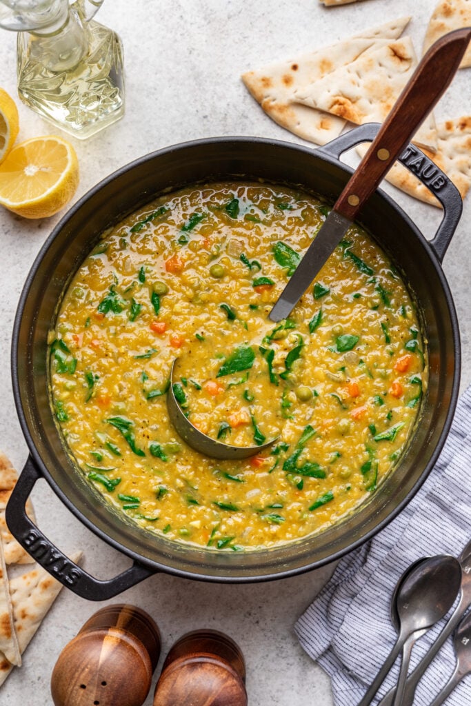 A pot of yellow lentil soup with spinach and carrots sits on a light countertop, surrounded by pita bread slices, lemon halves, a striped napkin, and pepper mills. A ladle rests inside the pot.