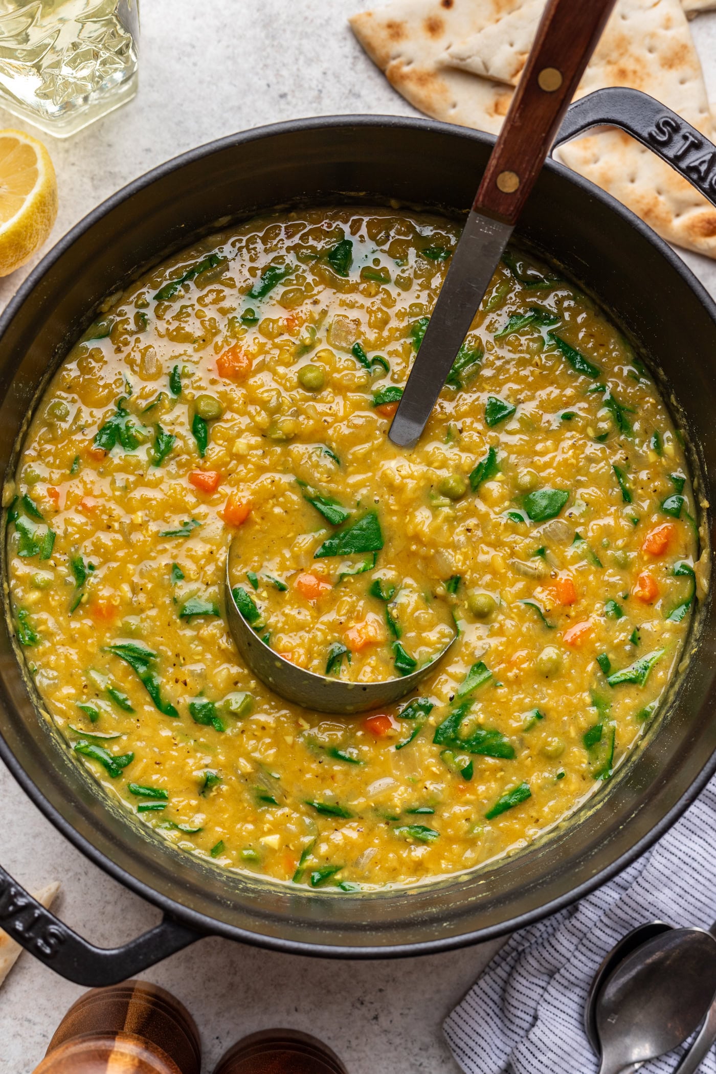 A pot of yellow lentil soup with chopped spinach and diced vegetables, stirred with a ladle. Naan bread, a lemon wedge, and utensils are visible around the pot on a light countertop.