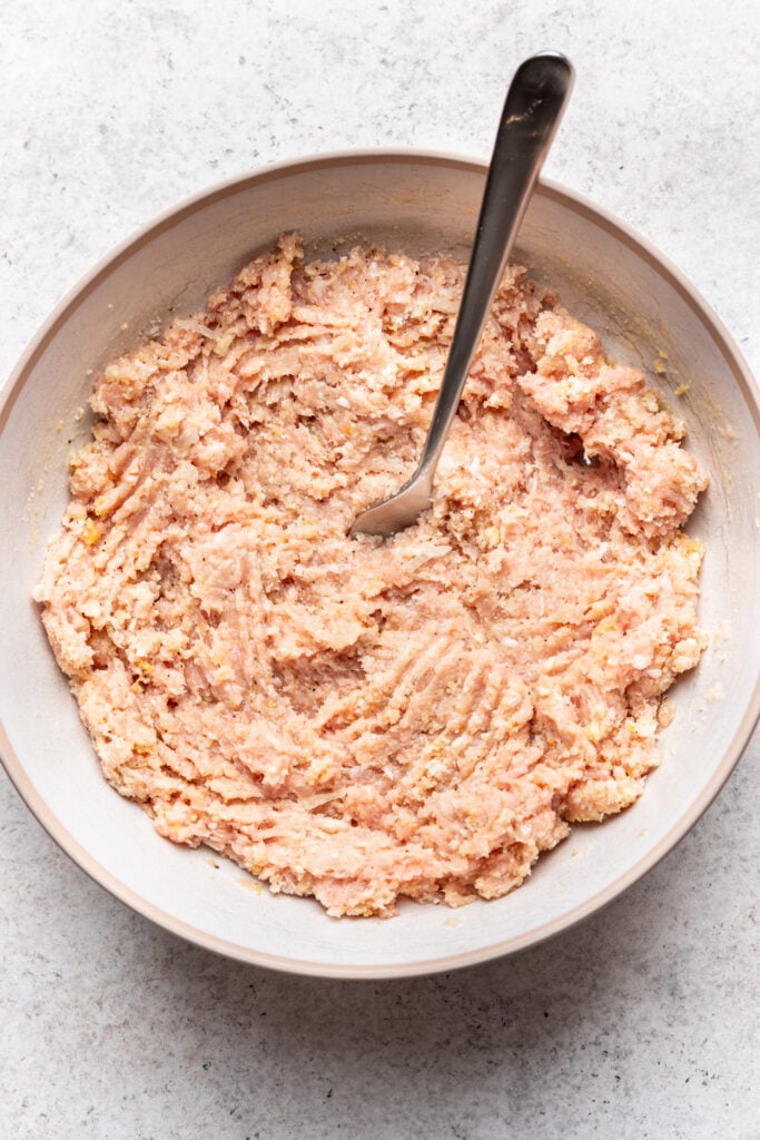 A mixing bowl filled with raw ground meat mixture and a spoon resting inside. The mixture appears to be evenly blended and has a pale pink color, likely prepared for cooking.