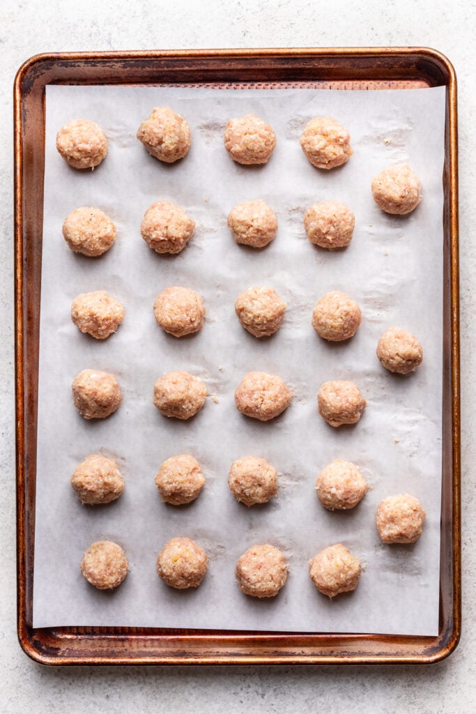 A baking sheet lined with parchment paper holds evenly spaced rows of uncooked meatballs, ready to be baked.