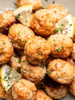 A close-up of golden-brown meatballs garnished with chopped parsley, grated cheese, and lemon wedges on a plate.