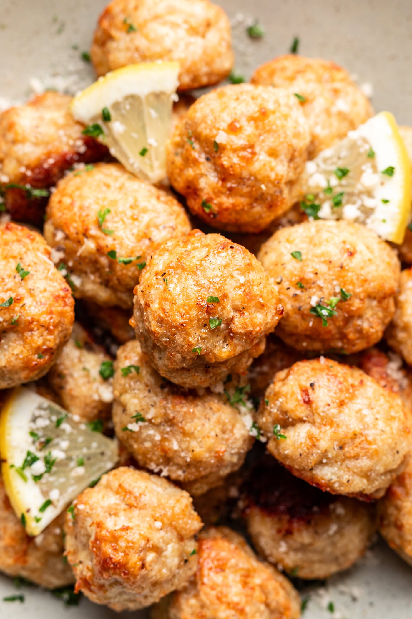 A close-up of golden-brown meatballs garnished with chopped parsley, grated cheese, and lemon wedges on a plate.
