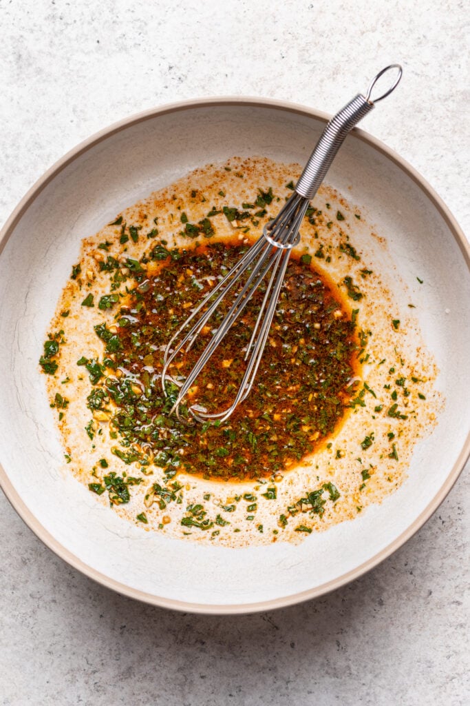 A metal whisk rests in a beige bowl containing a mixed marinade of oil, herbs, and spices on a light surface.