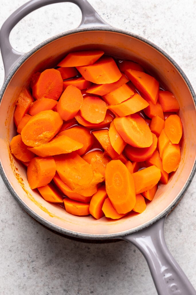 A gray pot filled with sliced, cooked carrots on a light-colored countertop. The carrots are bright orange and appear tender.