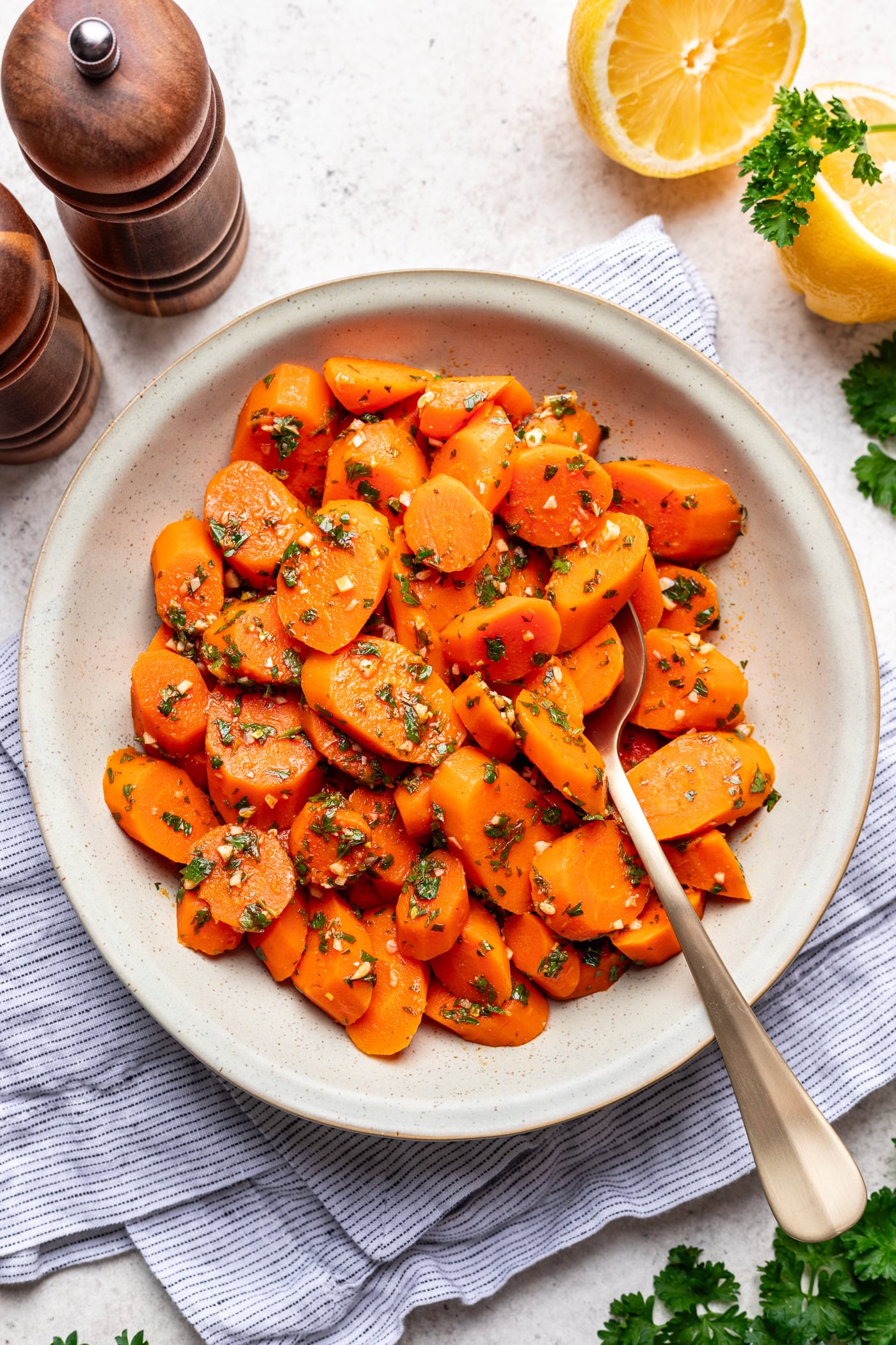 A bowl of sliced cooked carrots garnished with chopped herbs sits on a striped cloth, with a gold spoon, pepper mills, lemon halves, and parsley nearby.