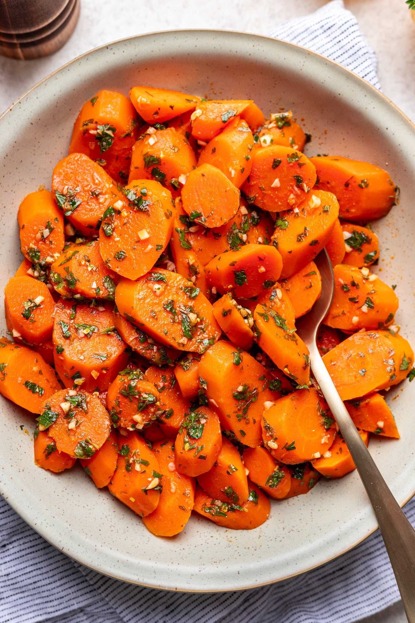 A bowl of sliced cooked carrots garnished with chopped herbs and minced garlic, served with a spoon on a light, striped cloth.