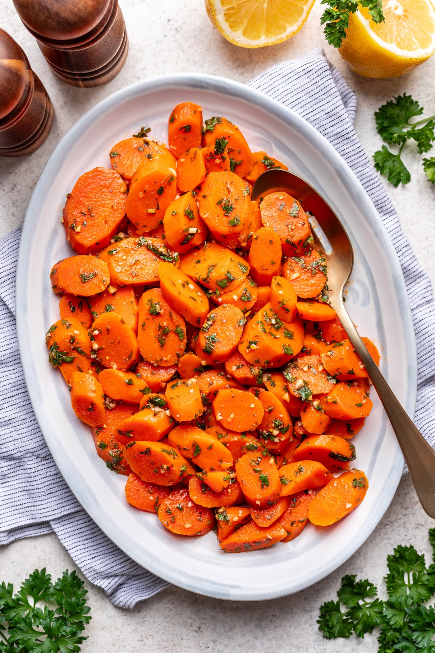 Oval white platter with sliced cooked carrots garnished with chopped parsley and herbs, served with a large serving spoon. Surrounding the dish are lemon halves, fresh parsley, salt and pepper shakers, and a striped cloth napkin.
