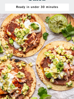 Three baked salmon tacos topped with avocado corn salsa and a drizzle of white sauce, served on corn tortillas with refried beans, garnished with cilantro and a halved lime on a light-colored surface.