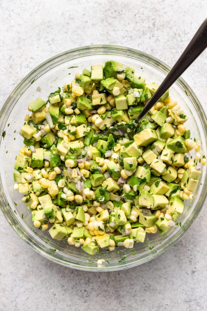 A glass bowl filled with a fresh salad of diced avocado, corn kernels, chopped herbs, with a spoon resting inside, on a light-colored surface.