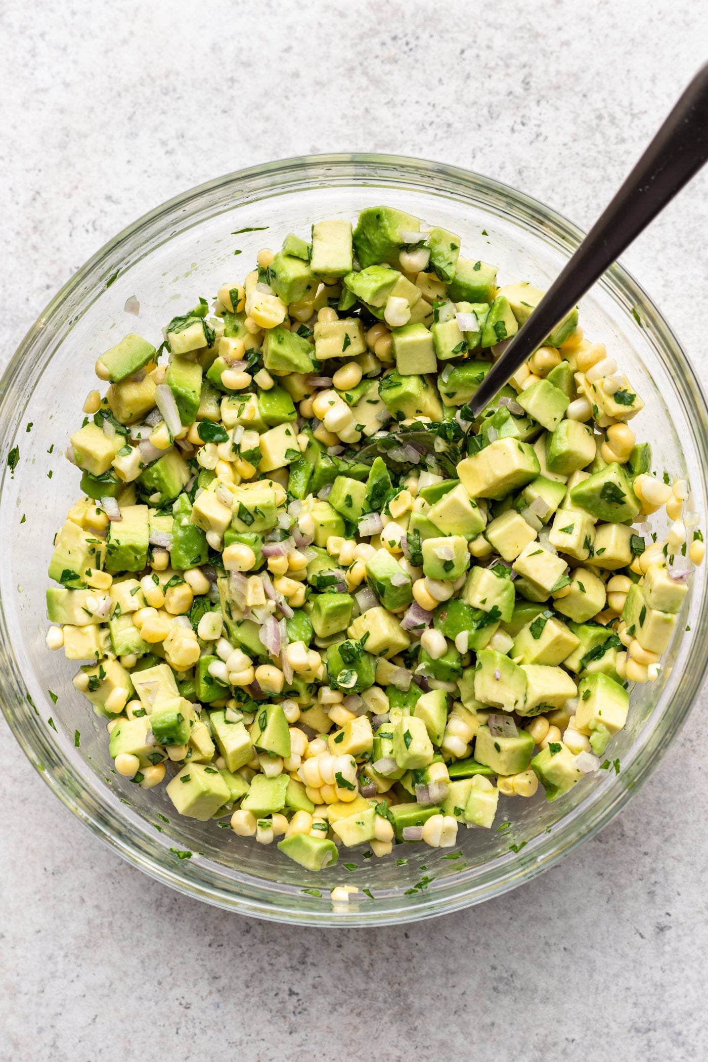 A glass bowl filled with a fresh salad of diced avocado, corn kernels, chopped herbs, with a spoon resting inside, on a light-colored surface.